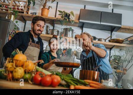 Lächelnde Köchin, die flammendes Essen in der Bratpfanne rührt Stehen bei Kollegen in der Küche Stockfoto