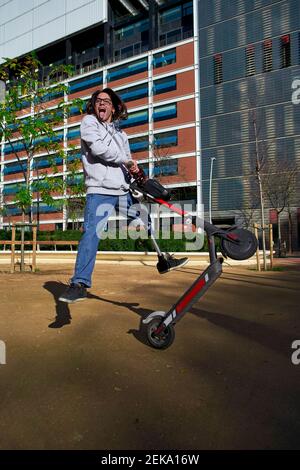 Fröhlicher junger behinderter Mann üben Stunt auf Elektro-Push-Roller Im Spielplatz gegen Gebäude Stockfoto