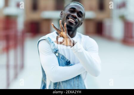 Lächelnder junger Mann in Denim Lätzchen Overalls gestikulierende OK Zeichen Stockfoto