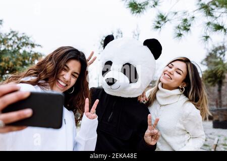Fröhliche weibliche Freunde, die Selfie mit einem männlichen Freund in Panda machen Maske während der Geste Friedensschild im Park Stockfoto