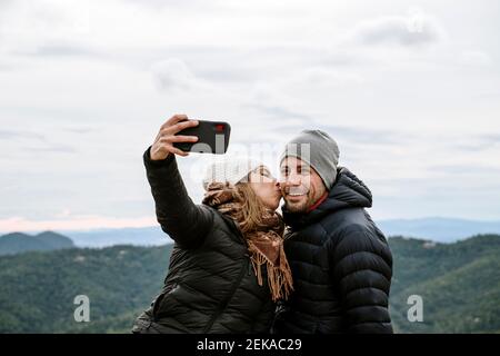 Frau küsst lächelnd Freund während Selfie gegen Himmel während Winter Stockfoto