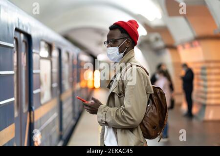 Schwarzer Mann trägt Gesichtsmaske als Schutz gegen covid-19 Virus, stehend in der U-Bahn, mit Handy. Stockfoto
