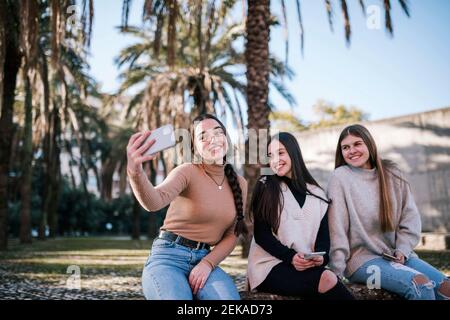 Lächelndes Teenager-Mädchen, das Selfie mit Freunden nimmt, während es dagegen sitzt Bäume im Park Stockfoto