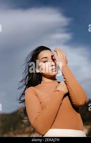 Junge Frau, die die Augen mit der Hand abschirmt, während sie bei Sonnenuntergang gegen den bewölkten Himmel steht Stockfoto