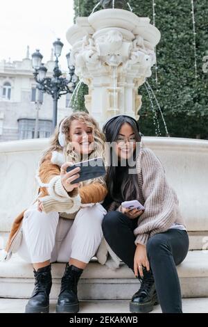 Multiethnische Freundinnen, die während des Sitzens Selfie über das Smartphone machen Gegen den Brunnen Stockfoto