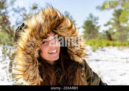 Lächelnde junge Frau trägt Fell Kapuzen Wintermantel starren während Im Wald stehen Stockfoto