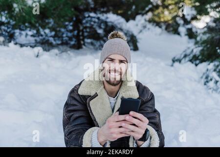 Lächelnder junger Mann, der im Schnee sitzt und das Handy hält Stockfoto