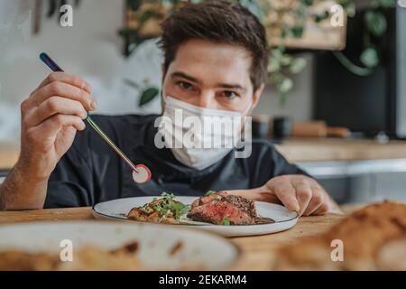 Männlicher Koch trägt schützende Gesichtsmaske garnieren Gemüse auf tomahawk Steak während der Arbeit in der Küche Stockfoto