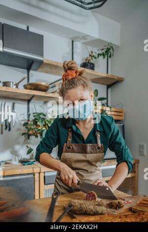 Junge Köchin trägt schützende Gesichtsmaske schneiden Salami während Arbeiten in der Küche während COVID-19 Stockfoto