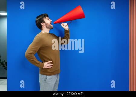 Geschäftsmann mit Hand auf Hüfte schreien in Megaphon von blau Wand am Arbeitsplatz Stockfoto