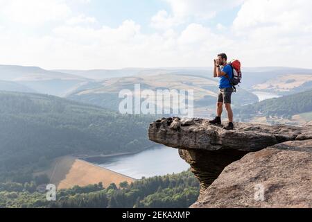 Junger männlicher Wanderer, der durch ein Fernglas schaut, während er auf einer Klippe steht Während der Ferien Stockfoto