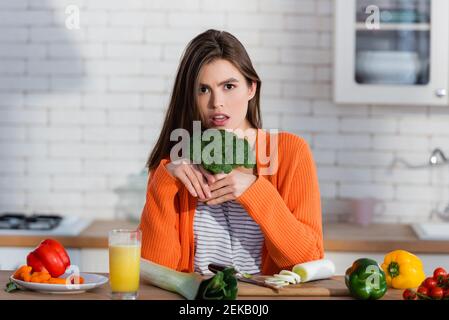 Seriöse Frau, die die Kamera anschaut, während sie Brokkoli nahe frisch hält Gemüse auf dem Tisch Stockfoto