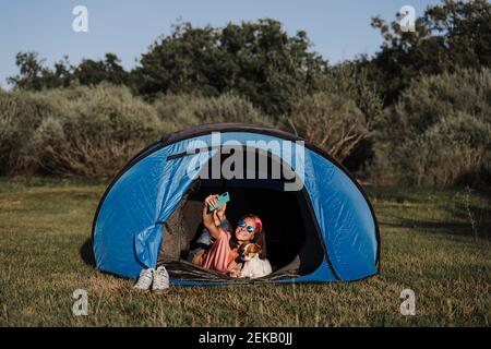 Frau trägt eine Sonnenbrille und nimmt ein Selfie mit dem Hund auf, während sie darin liegt Zelt Stockfoto
