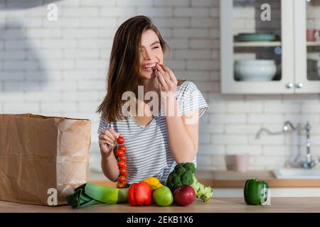 Lachende Frau beim Essen von Kirschtomaten in der Nähe von Äpfeln und frischem Gemüse In der Küche Stockfoto