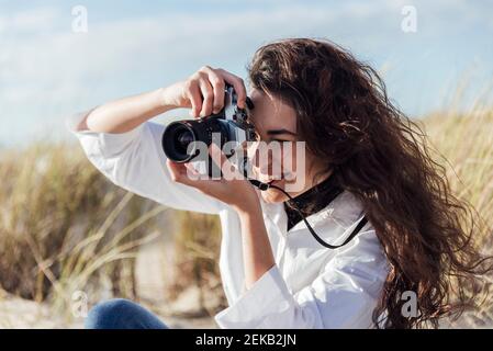 Junge Frau mit langen Haaren fotografiert durch Vintage-Kamera an Strand an sonnigen Tagen Stockfoto