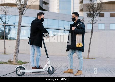 Junger Mann mit Elektroroller im Gespräch mit männlichen Freund tragen Beutel mit Zitronen während der Pandemie gegen Gebäude stehen Stockfoto