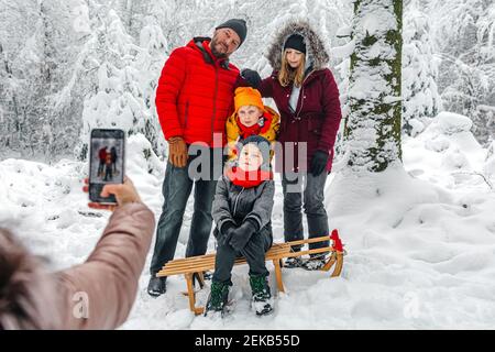 Frau, die im Stehen mit dem Mobiltelefon die Familie fotografiert Im Wald im Winter Stockfoto