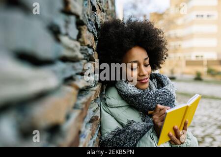 Lächelnde Afro-Frau, die in einem Buch schreibt, während sie sich auf Stein stützt Wand im Winter Stockfoto