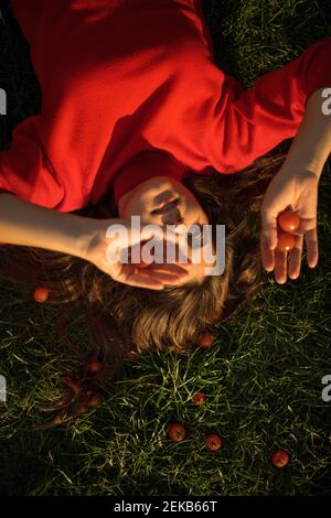 Junge Frau in rotem Kleid hält Kirschtomaten während liegend Auf Gras an sonnigen Tagen Stockfoto