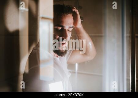 Junger Mann mit geschlossenen Augen unter der Dusche durch Glas gesehen Im Badezimmer Stockfoto