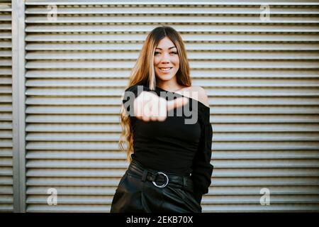 Lächelnde junge Frau, die Daumen nach oben zeigt, während sie gegen die Wand steht Stockfoto