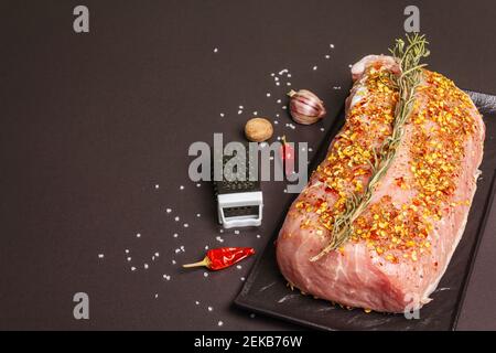 Rohe Schweinelende. Frisches Fleisch mit Rosmarin, Knoblauch und Flocken Gewürze auf dem Keramikständer. Reibe, getrocknete Chili, Salz, schwarzer Stein Beton Hintergrund Stockfoto