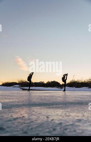 Zwei weibliche Eiskunstläuferinnen üben zusammen auf gefrorenem See bei Abenddämmerung Stockfoto