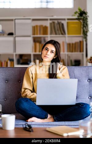 Junge Frau starrt, während sie mit Laptop zu Hause sitzt Stockfoto