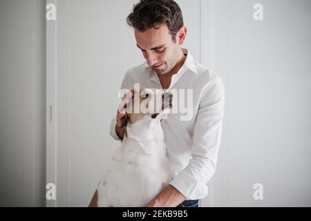 Mittelalter Mann streichelender Hund, während er gegen die weiße Wand steht Zu Hause Stockfoto
