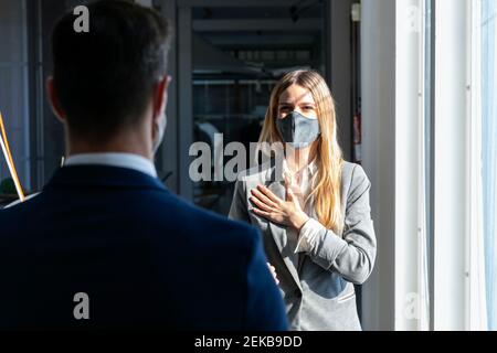 Geschäftsfrau trägt schützende Gesichtsmaske stehen mit der Hand auf der Brust Während der Begrüßung des Kollegen im Büro Stockfoto