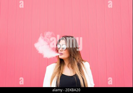 Junge Frau in einer Sonnenbrille atmet Rauch an der rosa Wand aus Stockfoto