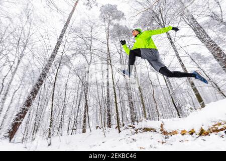 Männlicher Athlet, der über schneebedeckten gefallenen Baum im Wald springt Im Winter Stockfoto