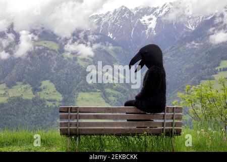 Frau im Krähenkostüm sitzt auf der Bank vor Bergkette Stockfoto
