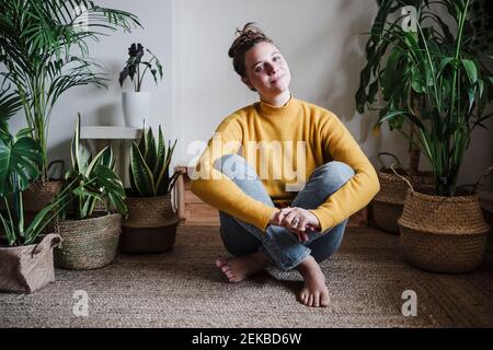 Junge Frau umarmt Knie, während sie zu Hause auf dem Teppich sitzt Stockfoto