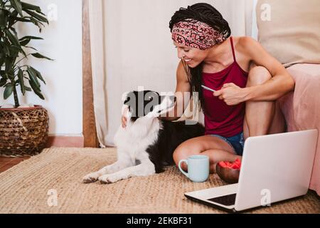 Lächelnde Frau mit Handy, die beim Sitzen auf den Hund schaut Mit dem Laptop auf dem Teppich zu Hause Stockfoto