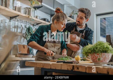 Der junge Koch riecht Petersilie, während er die beim Kollegen stehenden Frühlingszwiebeln schneidet Auf Kücheninsel Stockfoto