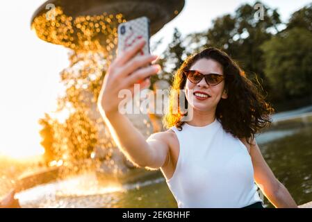 Junge Frau trägt eine Sonnenbrille und nimmt Selfie mit dem Smartphone entgegen Brunnen im Park Stockfoto