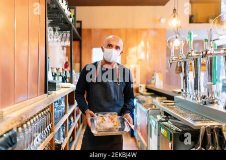 Reifer männlicher Besitzer, der die Lebensmittelbehälter in der Bar aushält Während COVID-19 Stockfoto