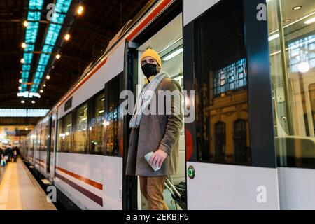 Mittelerwachsener Mann mit Gesichtsmaske beim Aussteigen aus dem Zug Mit Kaffeetasse und Gepäck am Bahnhof Stockfoto