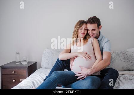 Lächelnder Mann berührt den Magen der schwangeren Frau, während er auf dem Bett sitzt Zu Hause Stockfoto