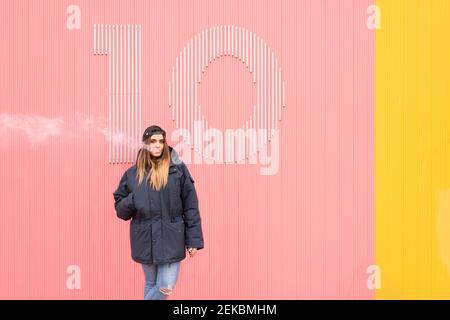 Junge Frau in Jacke atmet Rauch an rosa und gelben Wänden aus Stockfoto