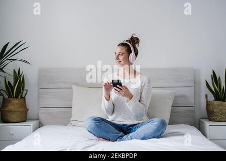 Junge Frau, die Kopfhörer trägt, während sie auf dem Mobiltelefon sitzt Bett zu Hause Stockfoto