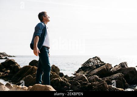 Genießen Sie reifen Mann stehen inmitten Felsen am Meer während sonnig Tag Stockfoto