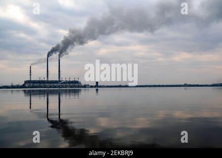 Kohlekraftwerk hohe Rohre mit schwarzem Rauch, der nach oben die verschmutzende Atmosphäre mit den Reflexionen von ihm im Seewasser bewegt. Stockfoto