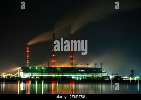 Hell erleuchtetes Kohlekraftwerk hohe Rohre mit schwarzem Rauch, der nach oben die verschmutzende Atmosphäre in der Nacht mit den Reflexen von ihm im Seewasser bewegt. Stockfoto