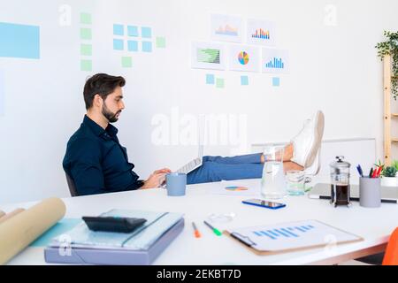 Männlicher Unternehmer, der am Laptop mit den Füßen am Schreibtisch arbeitet Gegen die Wand im Büro Stockfoto