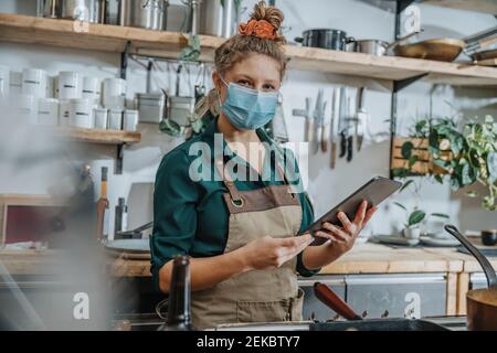 Weibliche Köchin trägt Schutzmaske mit digitalen Tablet während Stehen in der Küche Stockfoto