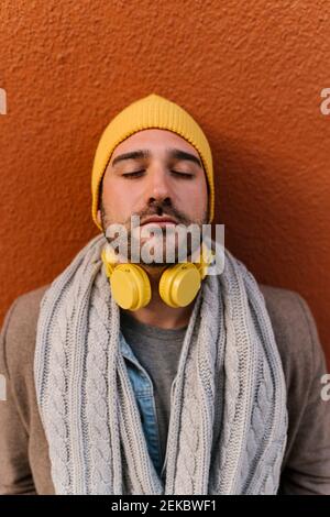 Mann trägt Strickmütze und Kopfhörer stehen mit geschlossenen Augen Gegen die Wand Stockfoto