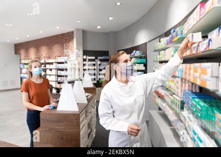 Der Kunde steht an der Kasse und schaut sich die Apothekerin an, die Medizin nimmt Aus dem Regal im Laden Stockfoto