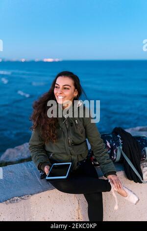 Schöne Frau mit digitalen Tablet sitzt auf Stützwand an Strand in der Dämmerung gegen Himmel Stockfoto